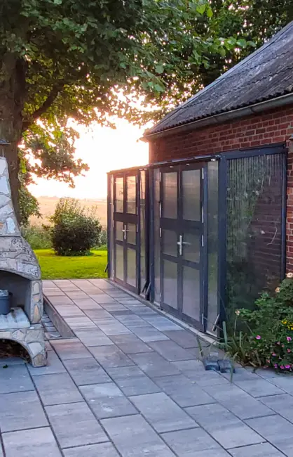 Anlehn-Tomatenhaus Modell TH-A 13 Version 2 mit Doppeltür in RAL 7016 Anthrazitgrau und Stegplatten an roter Backsteinwand bei Sonnenuntergang im Garten