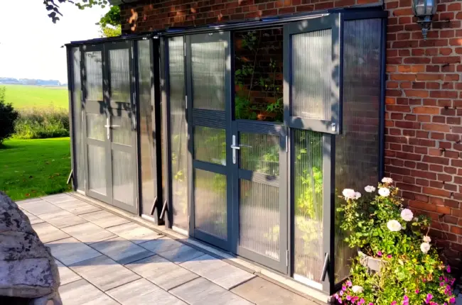 Anlehn-Tomatenhaus Modell TH-A 13 Version 2 mit Doppeltür in RAL 7016 und Stegplatten an Klinkerfassade, umgeben von gepflegtem Garten und Blühpflanzen