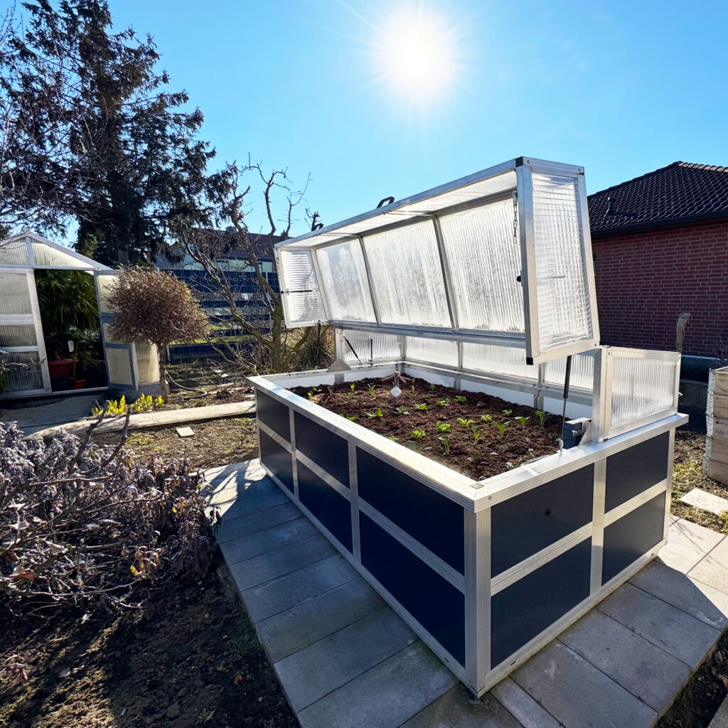 Hochbeet aus Aluminium mit anthrazitfarbenen Sandwichplatten und geöffnetem Frühbeet-Aufsatz bei direkter Sonneneinstrahlung im Garten