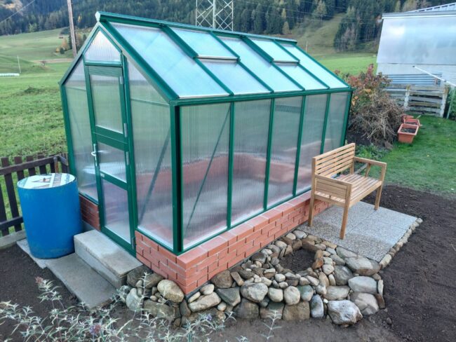 Kompaktes Gewächshaus aus grünem Aluminium mit Doppelstegplatten auf gemauertem Sockel im Garten, seitlicher Eingang und Regenfass daneben