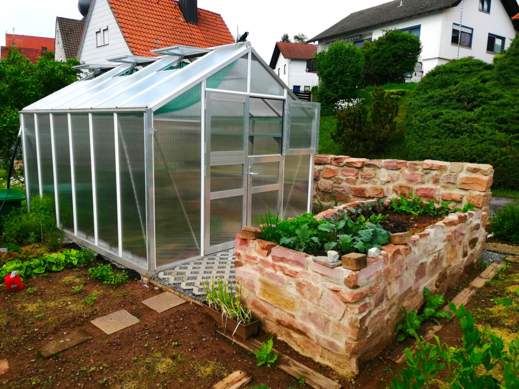 Gewächshaus mit Aluminium-Fundamentrahmen auf Punktfundamenten in einem privaten Garten mit Hochbeet aus Naturstein und geöffneter Tür.