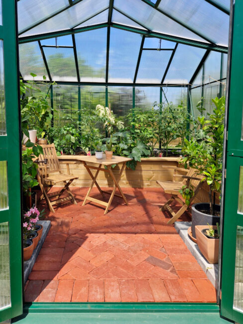 Der Innenraum eines grünen Gewächshauses mit transparenten Stegplatten. Auf dem roten Ziegelboden stehen Pflanzen in Töpfen sowie ein kleiner Holztisch mit zwei Stühlen. Das Gewächshaus ist dicht bepflanzt und hell.