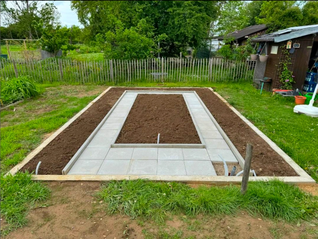 Vorbereitetes Streifenfundament für ein Gewächshaus mit Beetaufteilung sowie Leerrohren für Strom und Wasser.